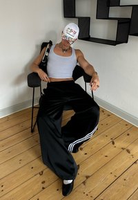 Juodos satino plačios kojinės kelnės su baltomis juostelėmis, derinamos su pilku marškinėliu be rankovių ir gėlių tematikos beisbolo kepuraite. Sėdi juodame kėdėje.