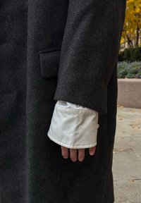 Manteau en laine gris foncé avec un poignet en satin blanc visible sous la manche. Le contraste de textures entre les matériaux est remarquable.