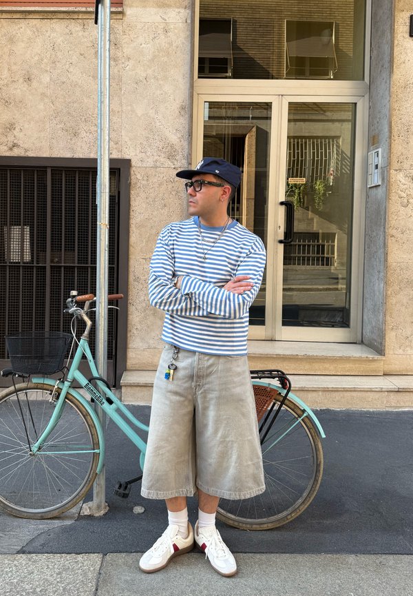 Hombre con camiseta de manga larga a rayas, pantalones cortos beige, gorra y gafas de sol, de pie con los brazos cruzados junto a una bicicleta verde menta en una acera de la ciudad.