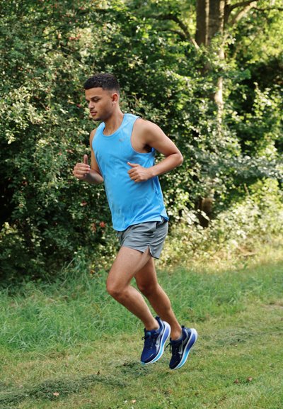 Corredor masculino atlético en una camiseta sin mangas azul y pantalones cortos grises, que lleva zapatillas de correr azules, trotando por un sendero de hierba rodeado de vegetación.