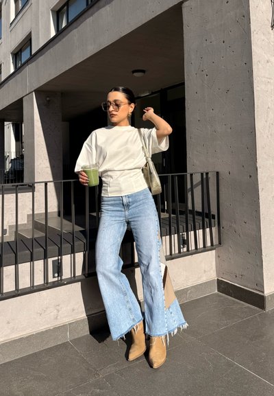 Mujer con top blanco, vaqueros anchos de pierna en azul claro, botas marrones y gafas de sol, sosteniendo una bebida verde, de pie junto a una barandilla en un entorno urbano soleado.