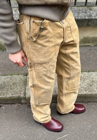 Άτομο με μπεζ παντελόνι cargo, με αλυσίδα ως αξεσουάρ, και μπορντό δερμάτινα παπούτσια, στέκεται σε πεζοδρόμιο κοντά σε σκαλοπάτια από πέτρα.