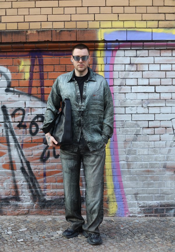 Egy férfi kopott farmer ruhában és napszemüvegben, fekete táskát tartva, graffitivel borított téglafal előtt áll.