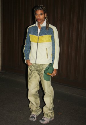 Jacket with blue denim, white, and yellow panels; zip closure. Olive green pants with a textured pattern. Green cap with orange text. Sneakers.