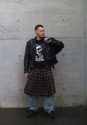 Hombre con chaqueta de cuero negra, camiseta con un gráfico de calavera, falda escocesa a cuadros sobre vaqueros holgados y zapatillas negras, de pie contra una pared de hormigón.