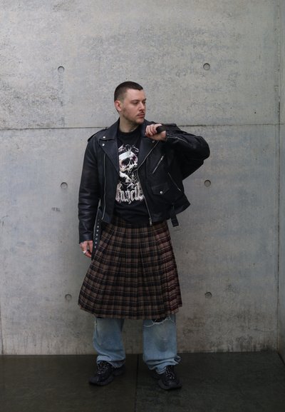 Hombre con chaqueta de cuero negra, camiseta con un gráfico de calavera, falda escocesa a cuadros sobre vaqueros holgados y zapatillas negras, de pie contra una pared de hormigón.