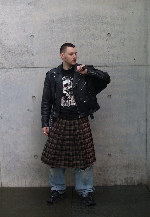 Uomo che indossa una giacca di pelle nera, una maglietta con grafica di teschio, un kilt a quadri sopra jeans larghi e sneakers nere, in piedi contro un muro di cemento.