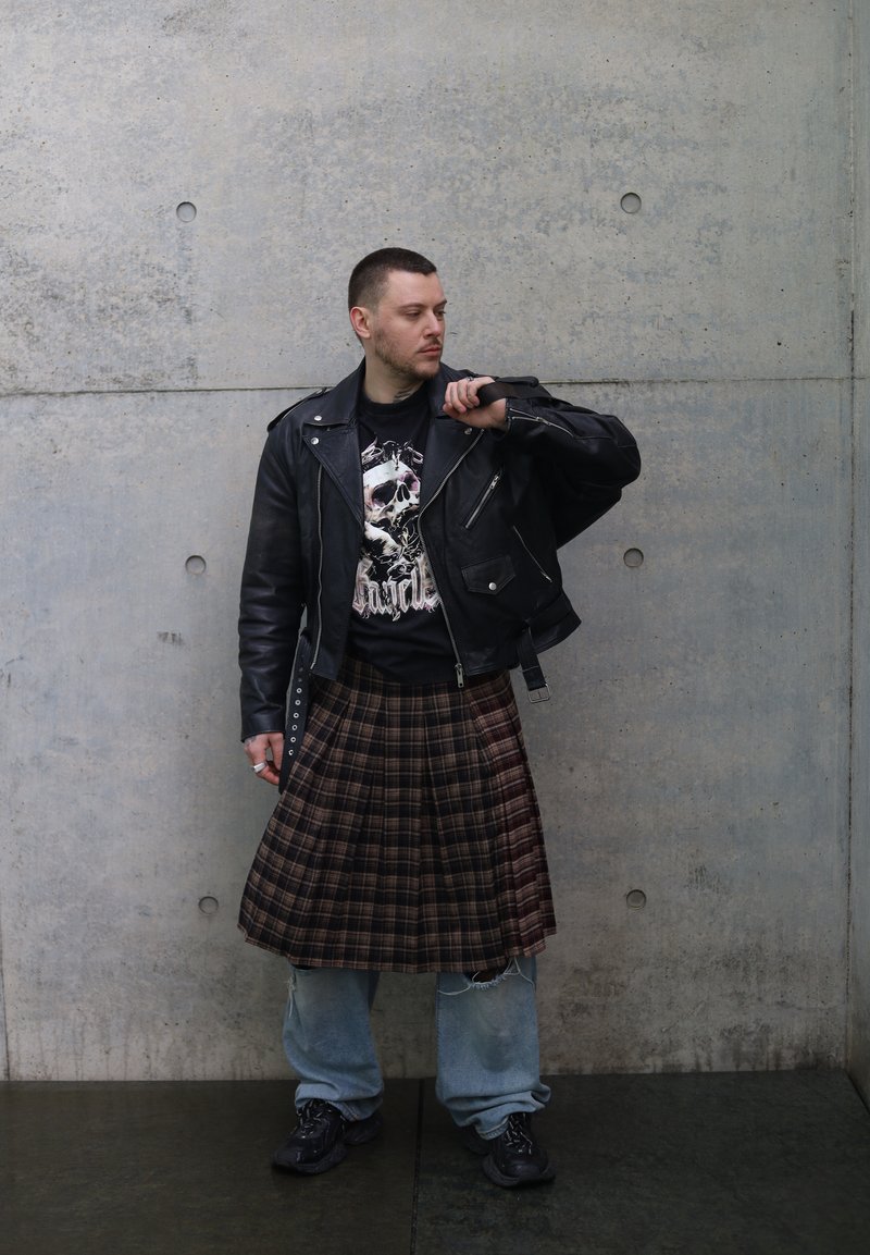 Muž v čiernej koženej bunde, tričku s lebkovým motívom, károvanom kiltu cez voľné rifle a čiernych teniskách stojí opretý o betónovú stenu.