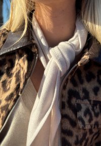 Écharpe blanche nouée en tissu doux, superposée sur un pull beige, associée à une veste à imprimé léopard avec une fermeture éclair.