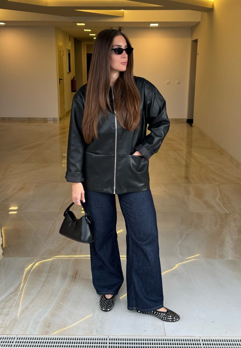 Veste en similicuir noir avec fermeture éclair à l'avant, pantalon en denim foncé évasé, ballerines noires cloutées, et petit sac à main noir. Cheveux longs et raides.