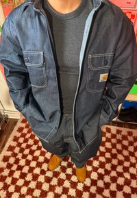 Jachetă din denim închis la culoare, cu două buzunare frontale, purtată peste un tricou negru cu dungi. Asortată cu pantaloni din denim de aceeași culoare și încălțăminte galben-muştar.