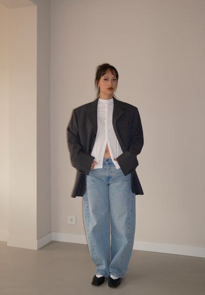 Blazer gris de gran tamaño sobre una camisa blanca de botones, combinado con jeans azules holgados y zapatos negros sin cordones, frente a un fondo neutro.