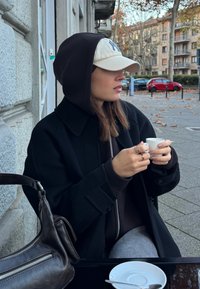 Črni plašč z velikim ovratnikom, rjava kapuca in svetla kapa. Drži belo skodelico; rjava torba počiva poleg nje na mizi.