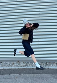 Τζην φούστα συνδυασμένη με μαύρο μπουφάν, γκρι σκούφο και μαύρα παπούτσια. Μικρή μπεζ τσάντα χεριού και λευκές κάλτσες ορατές. Φόντο με οριζόντιες ρίγες.