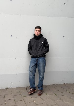 Chaqueta negra con capucha y cremalleras, jeans de mezclilla azules y zapatos marrones. De pie contra una pared suave y de color gris claro con pequeños detalles circulares.