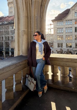 Mujer con gafas de sol, chaqueta oscura, bufanda azul y vaqueros apoyada en la barandilla de un balcón de piedra con vista a los edificios de la ciudad al atardecer.