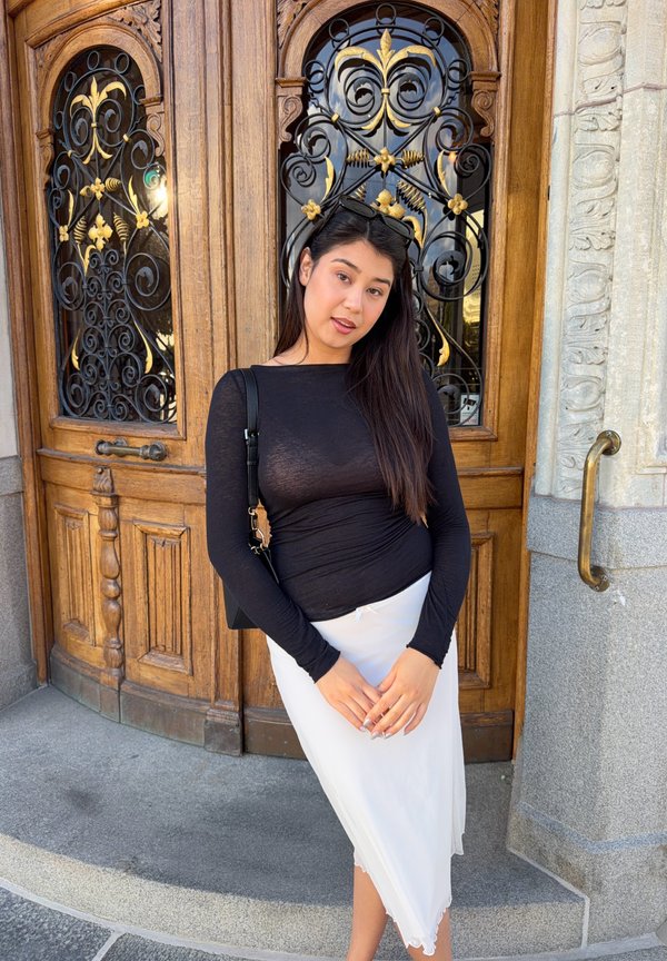Una mujer con un top negro y una falda blanca posa frente a una puerta de madera ornamentada con herrajes decorativos de hierro.