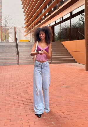 Modelo vistiendo un camisón lavanda con encaje, jeans de pierna ancha de lavado claro y zapatos negros, con las manos juntas en un entorno de patio.