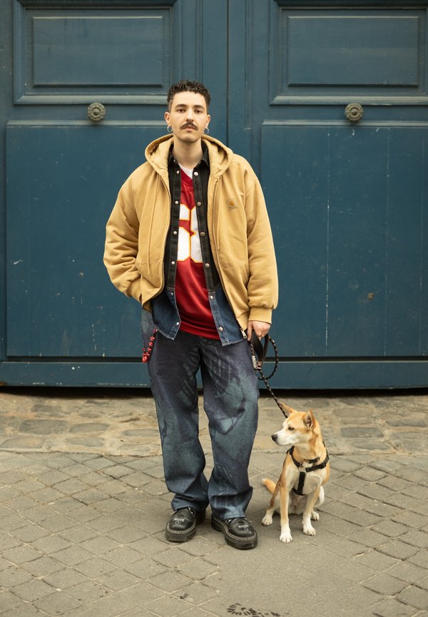 En person klädd i en beige hoodie över en svart tröja och en röd tröja, tillsammans med mörka jeans. En liten hund i koppel sitter bredvid dem.