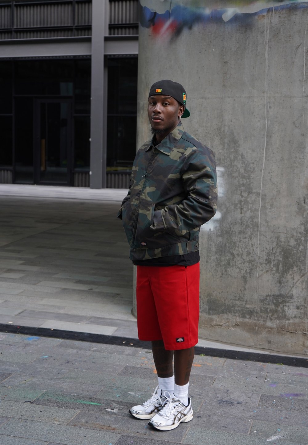Camouflagejas, rode shorts en zilveren sneakers. De setting heeft een betonnen muur met graffiti en een vlakke, verharde ondergrond.
