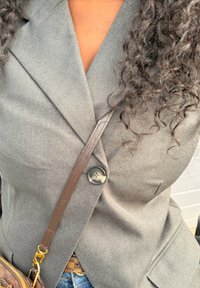 Άτομο που φοράει γκρι σακάκι με ένα κουμπί, σγουρά μαλλιά, μπλε τζιν και καφέ τσάντα χιαστί με χρυσά μεταλλικά στοιχεία.