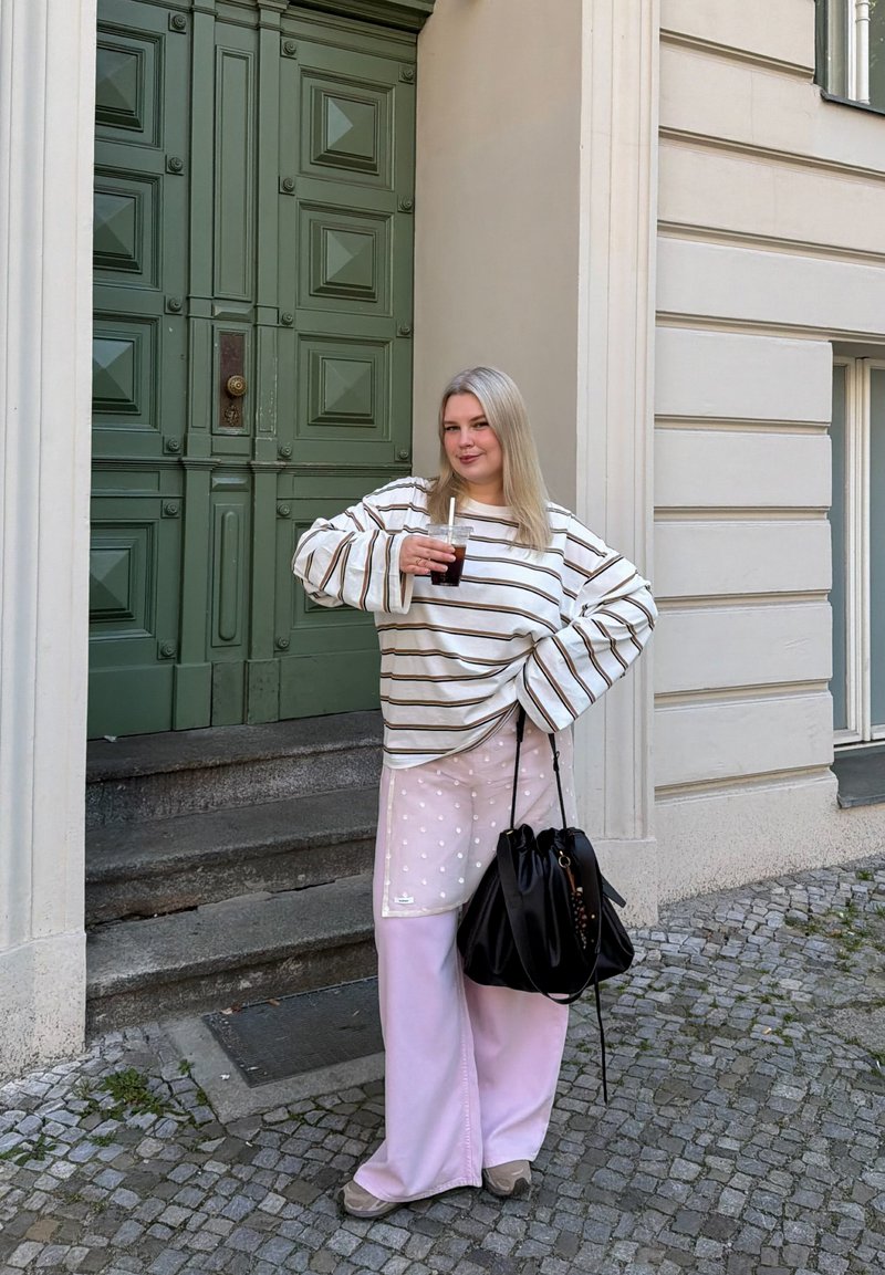 Jeune femme en chemise à manches longues rayée et pantalon rose tient une boisson et un sac noir, debout dans une rue pavée à côté d'une porte verte.