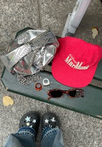 Stříbrná metalická taška s hroty, červená kordurová kšiltovka, dva prsteny (jeden oranžový, jeden bílý), brýle ve stylu želvoviny a zdobené černé dřeváky.