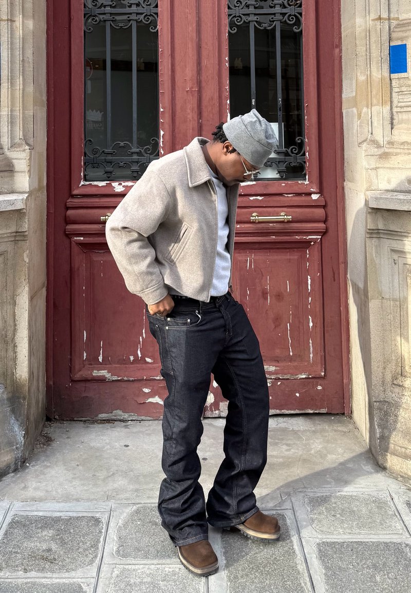 Homme portant une veste grise, un bonnet gris et un jean foncé, regardant vers le bas en se tenant sur un pavé en pierre devant des portes doubles rouges usées.