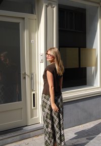Braunes Kurzarm-Oberteil kombiniert mit karierter, weiter Hose in Grün, Beige und Grau. Reflektierende Oberfläche und Türklingel sichtbar.
