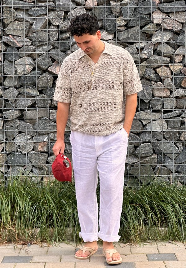 Homme aux cheveux bouclés et foncés, portant un pull beige en tricot, un pantalon en lin blanc et des sandales, se tient debout en tenant une casquette rouge devant un mur de pierre.