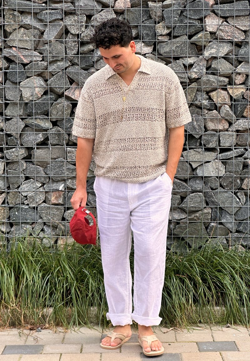 Mann mit lockigem dunklem Haar, der ein beiges Strickshirt, weiße Leinenhosen und Sandalen trägt, steht vor einer Steinmauer und hält eine rote Kappe.