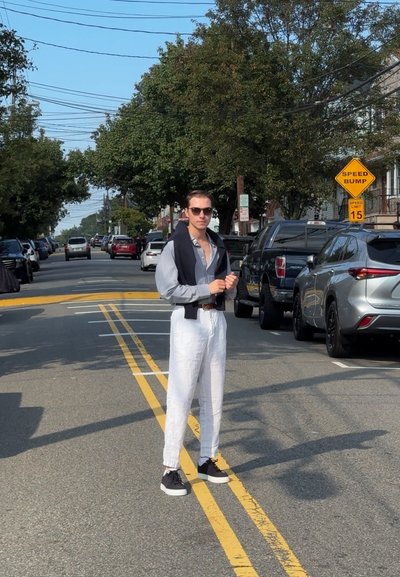 Pantalones de lino blancos y una camisa gris de botones llevada parcialmente desabrochada, con un suéter oscuro caído sobre los hombros. Zapatillas negras.
