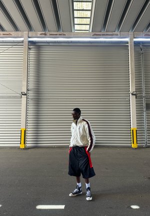 Hombre vestido con una chaqueta blanca y pantalones cortos negros, de pie con las manos en los bolsillos frente a una gran puerta persiana metálica industrial.