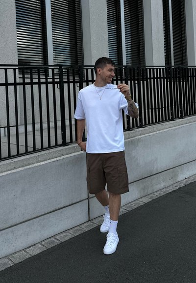 Camiseta blanca de manga corta, pantalones cortos de cargo marrones y zapatillas blancas. Cadena y pulsera de plata delgada. De pie en una acera cerca de un edificio.