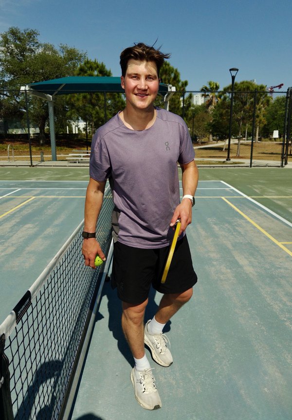 Joven con camiseta morada y pantalones cortos negros sosteniendo una pala y una pelota de pickleball en una cancha exterior soleada cerca de la red.