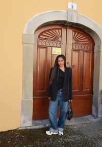 Blazer preto por cima de uma camisola preta, jeans azuis largos e sapatilhas claras. Em pé em frente a uma porta de madeira com elementos decorativos.