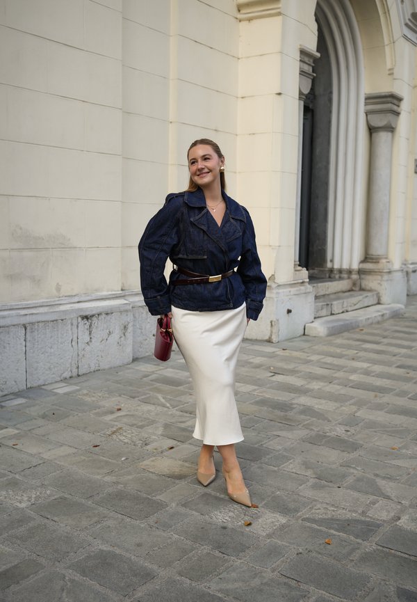 Jeansjacke mit großem Kragen und tailliertem Schnitt, kombiniert mit einem weißen Satinrock und nudefarbenen spitzen Schuhen, während sie eine bordeauxfarbene Handtasche hält.