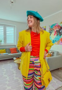 Helles gelbes Blazer über einem roten Hemd, Regenbogen-gestreifte Schlaghosen, türkiser Barett und eine gelbe Handtasche mit Kettenriemen.