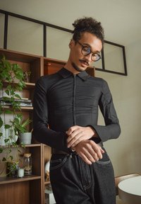 Zwart shirt met lange mouwen, een ritssluiting aan de voorkant en een kraag. Glad stof, aansluitend ontwerp en manchetten met knopen, gecombineerd met donkere spijkerbroek.