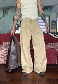 Person med tatoverte armer iført beige vide bukser og en hvit ermeløs strikket topp, som holder en mørkebrun lærjakke innendørs.
