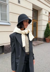 Čierny vlnený klobúk, krémový šál s pleteným vzorom, čierny oversize blejzer, béžová košeľa na gombíky, čierne džínsy, stojí vonku pri rastline.
