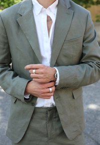 Chaqueta de traje de lino gris claro sobre una camisa blanca con botones, presentando múltiples anillos en las manos, con un reloj visible y una tela texturada.