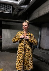 Γυναίκα με γυαλιά κουμπώνοντας ένα παλτό με ρίγες τίγρης σε ένα αμυδρά φωτισμένο γκαράζ στάθμευσης από σκυρόδεμα με εκτεθειμένους σωλήνες στην οροφή.