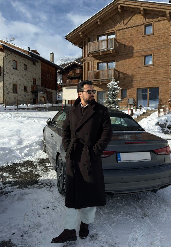 Brūns vilnas virsjaka ar jostu, gaiši dzelteni džinsa bikses un tumši brūni zābaki. Pelēks auto sniegotā kalnu ainavā, fonā koka ēkas.