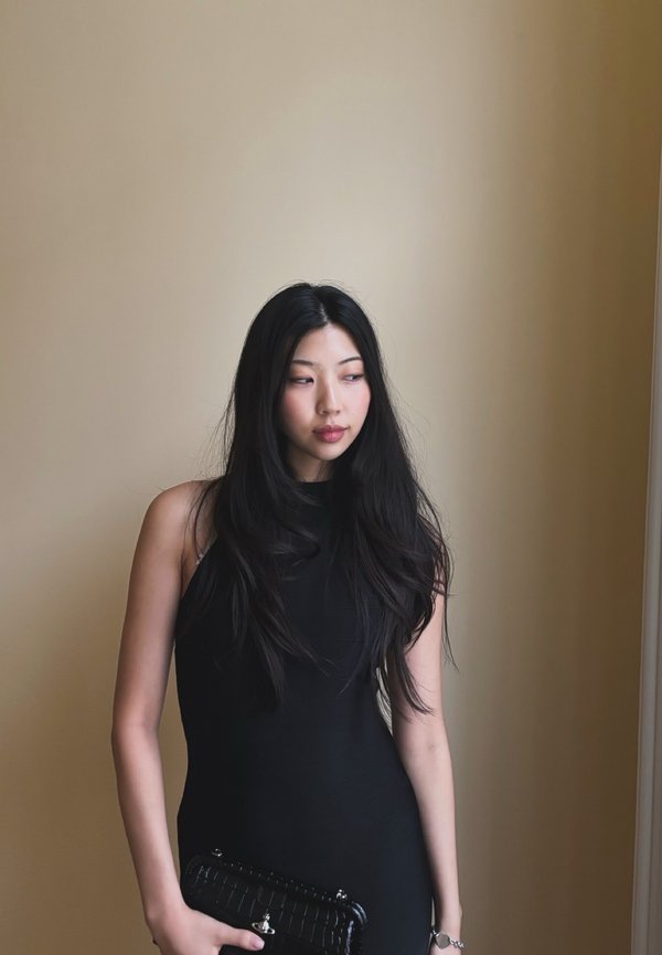 Jeune femme aux longs cheveux noirs portant une robe noire sans manches, tenant une pochette noire, debout contre un mur beige.