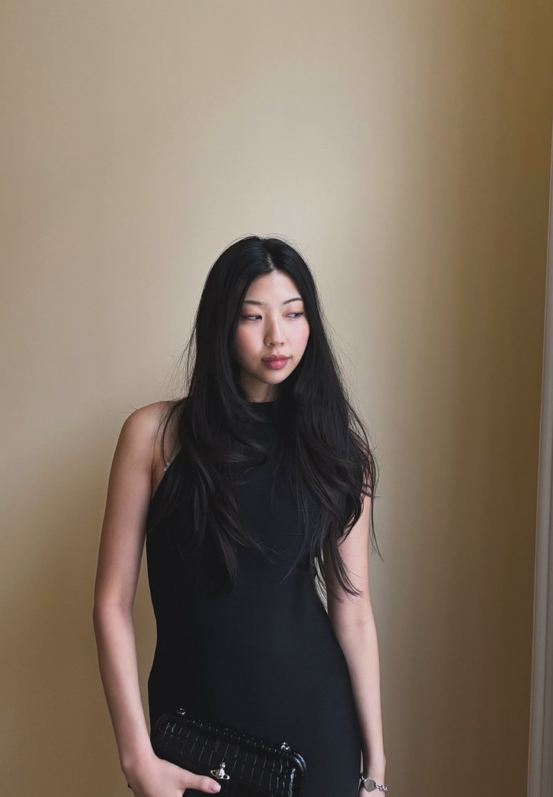 Jeune femme aux longs cheveux noirs portant une robe noire sans manches, tenant une pochette noire, debout contre un mur beige.