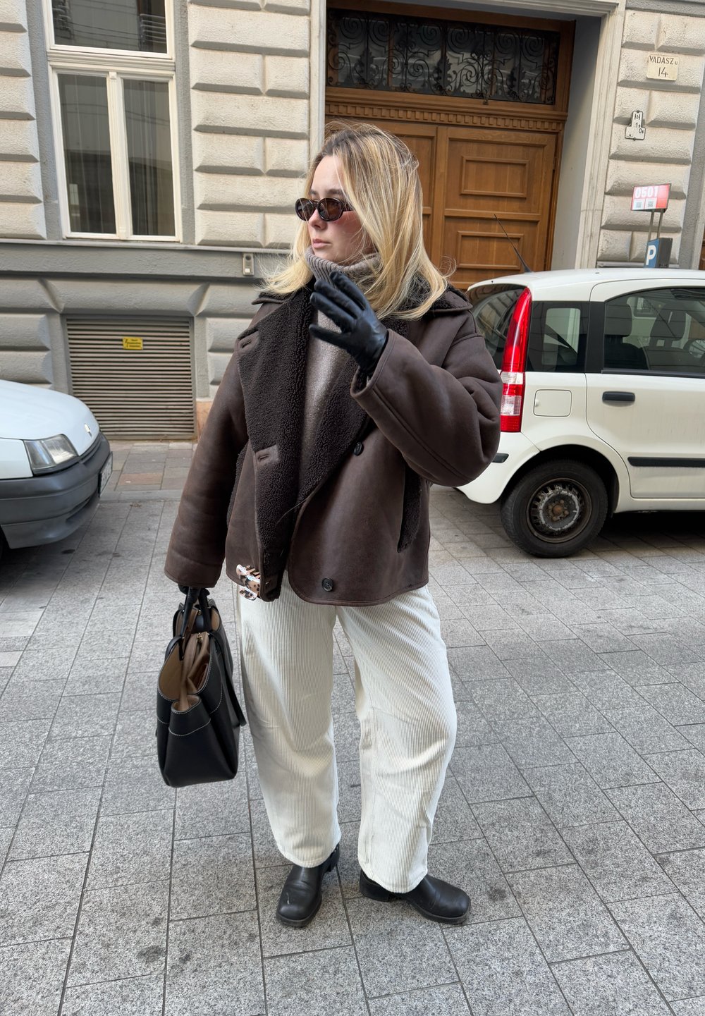 Καφέ παλτό με κολάρο από γούνα, λευκές ριγωτές παντελόνες, μαύρα δερμάτινα γάντια και μπότες, κρατώντας μια μαύρη τσάντα. Αστική σκηνή.