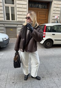 Καφέ παλτό με κολάρο από γούνα, λευκές ριγωτές παντελόνες, μαύρα δερμάτινα γάντια και μπότες, κρατώντας μια μαύρη τσάντα. Αστική σκηνή.