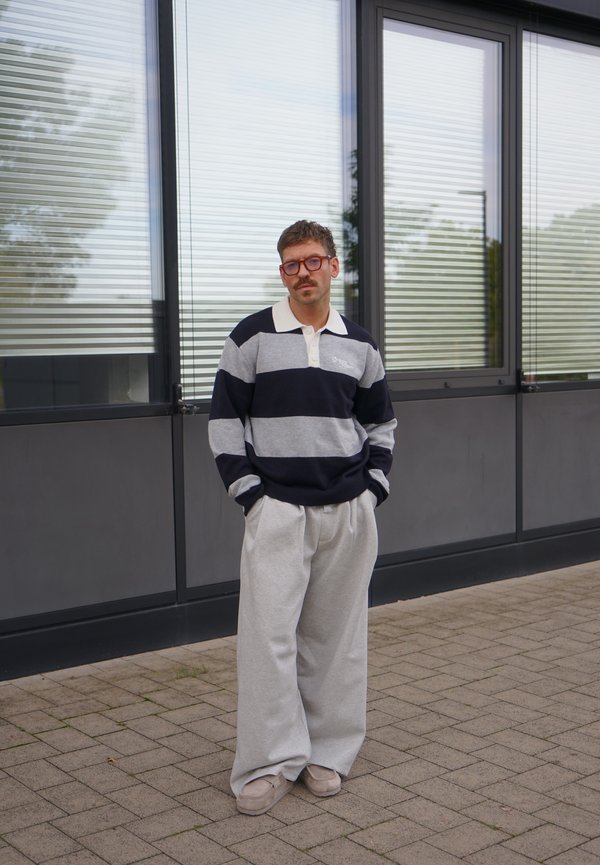 Hombre con gafas que lleva una camisa de manga larga a rayas negras y grises y pantalones anchos grises, de pie en una acera pavimentada frente a unas ventanas.