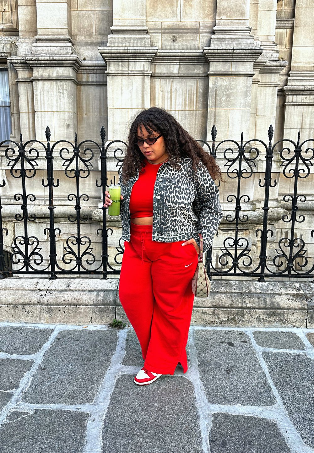 Red wide-leg trousers paired with a red crop top underneath a patterned grey leopard print jacket. Holding a green beverage, with a small bag and patterned trainers.